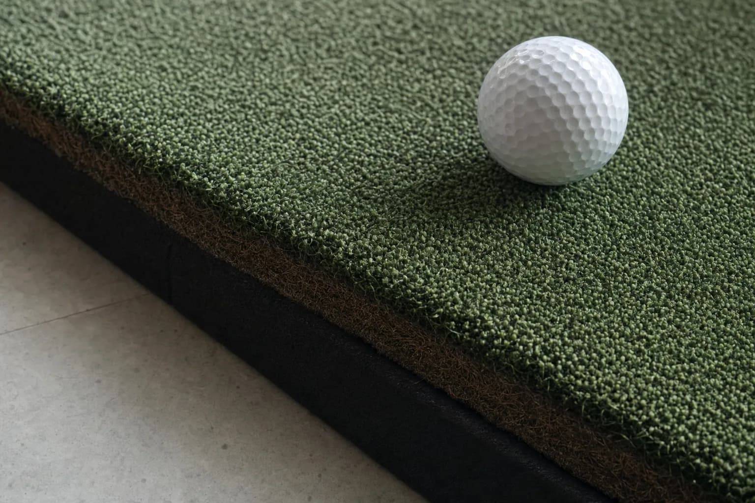 Close-up of layered artificial golf turf on a thick foam base at the edge of a hitting mat, a white golf ball on the surface