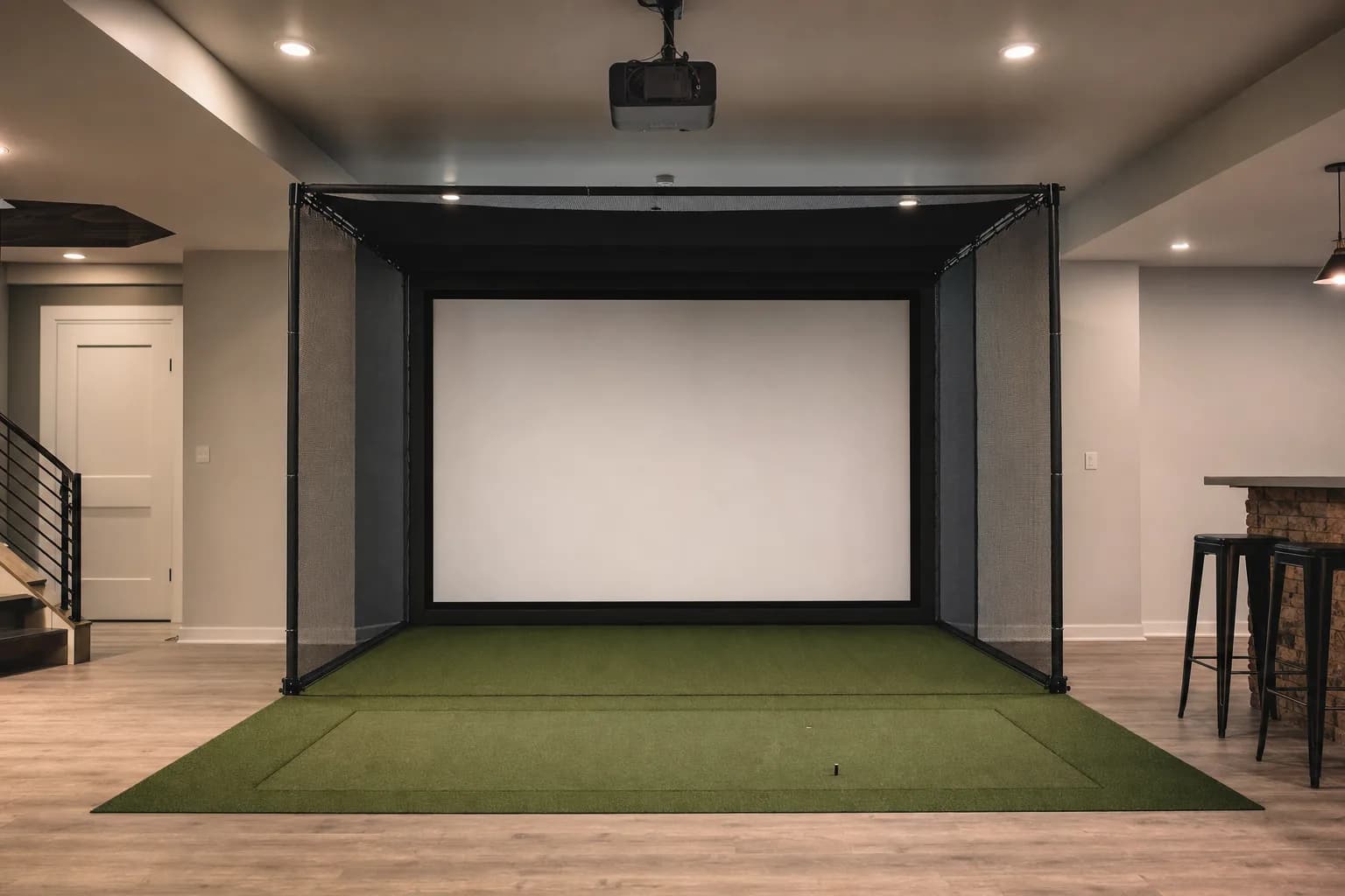 A complete but unoccupied home golf simulator in a finished basement: enclosure frame, impact screen, turf mat, side netting and a ceiling projector