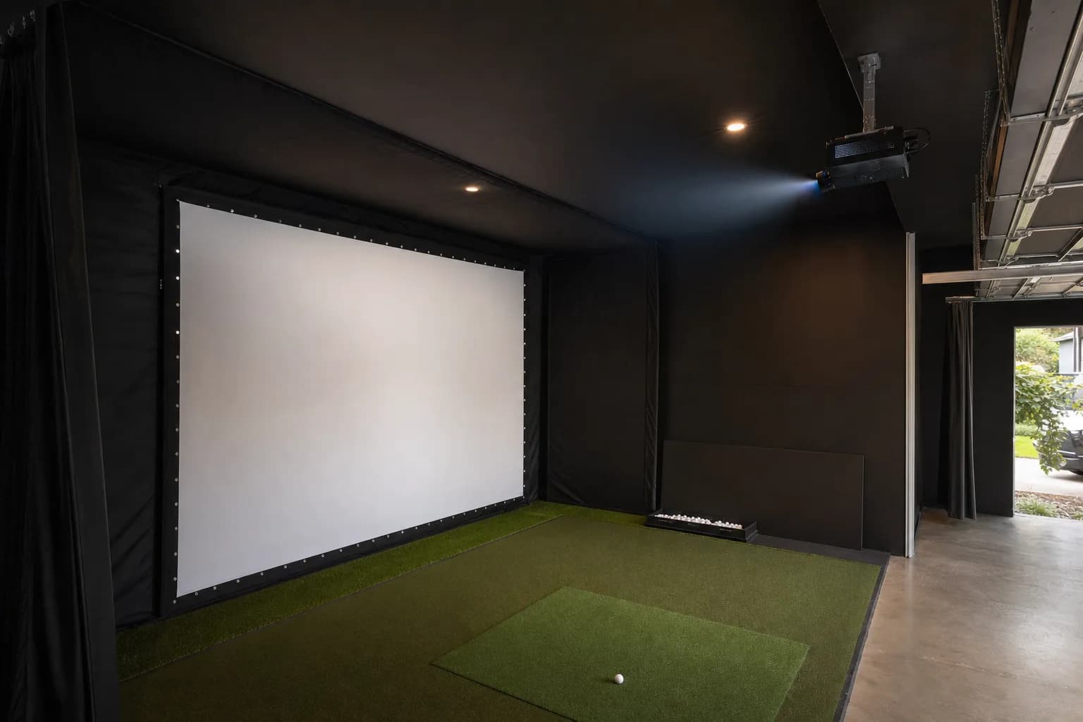 A tensioned golf simulator impact screen lit by a ceiling-mounted projector in a dark garage bay